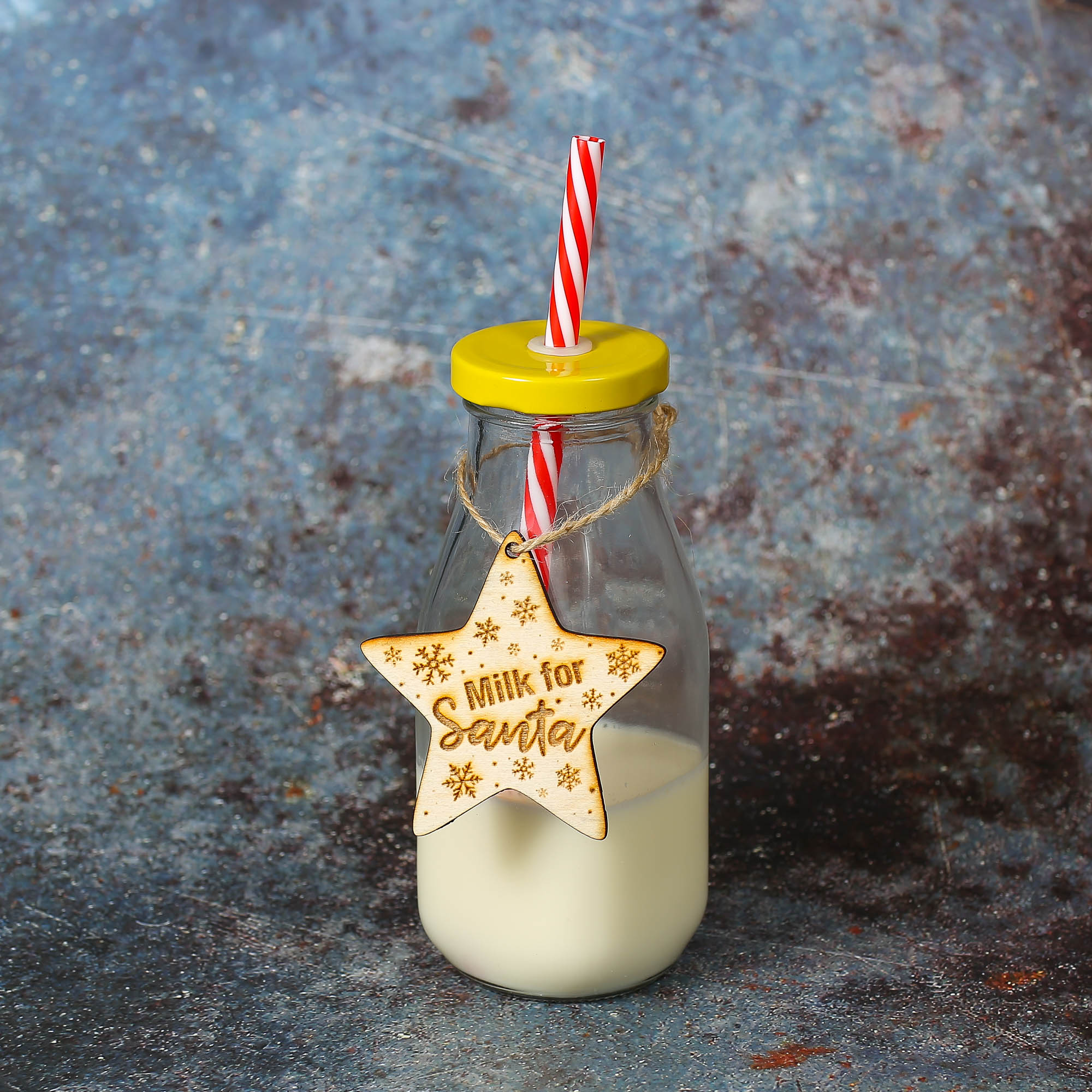 Glass bottle with yellow lid with straw and a wooden star-shaped decoration hanging from the bottle neck with milk for Santa engraved on it