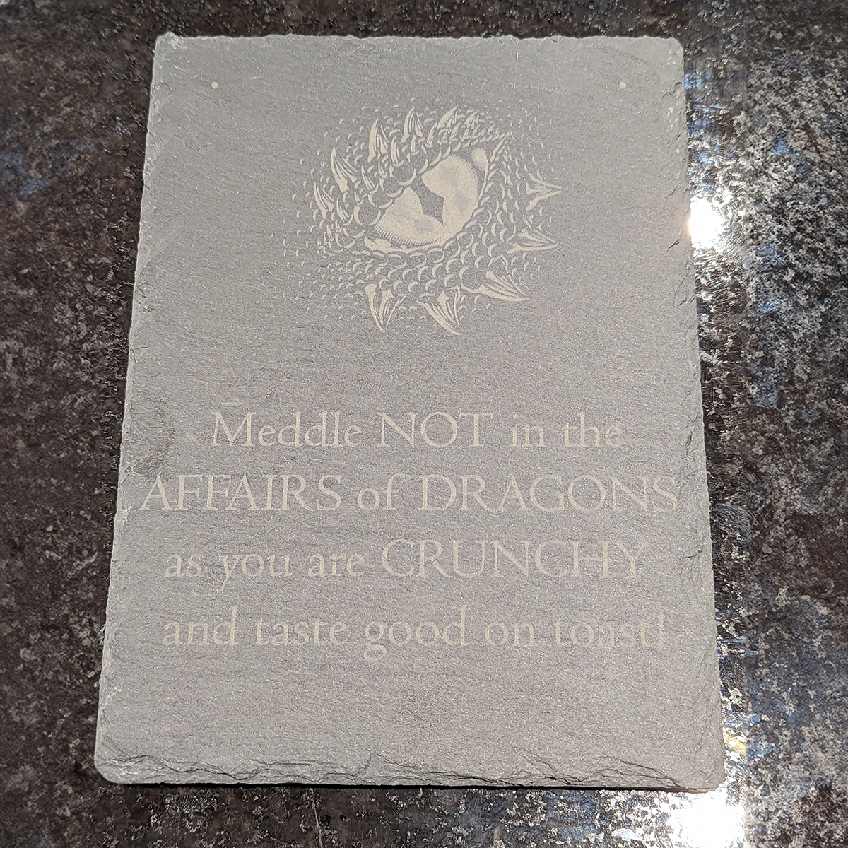 Slate sign with a dragon's eye and text saying Meddle not in the affairs of dragons as you are crunchy and taste good on toast engraved on it.