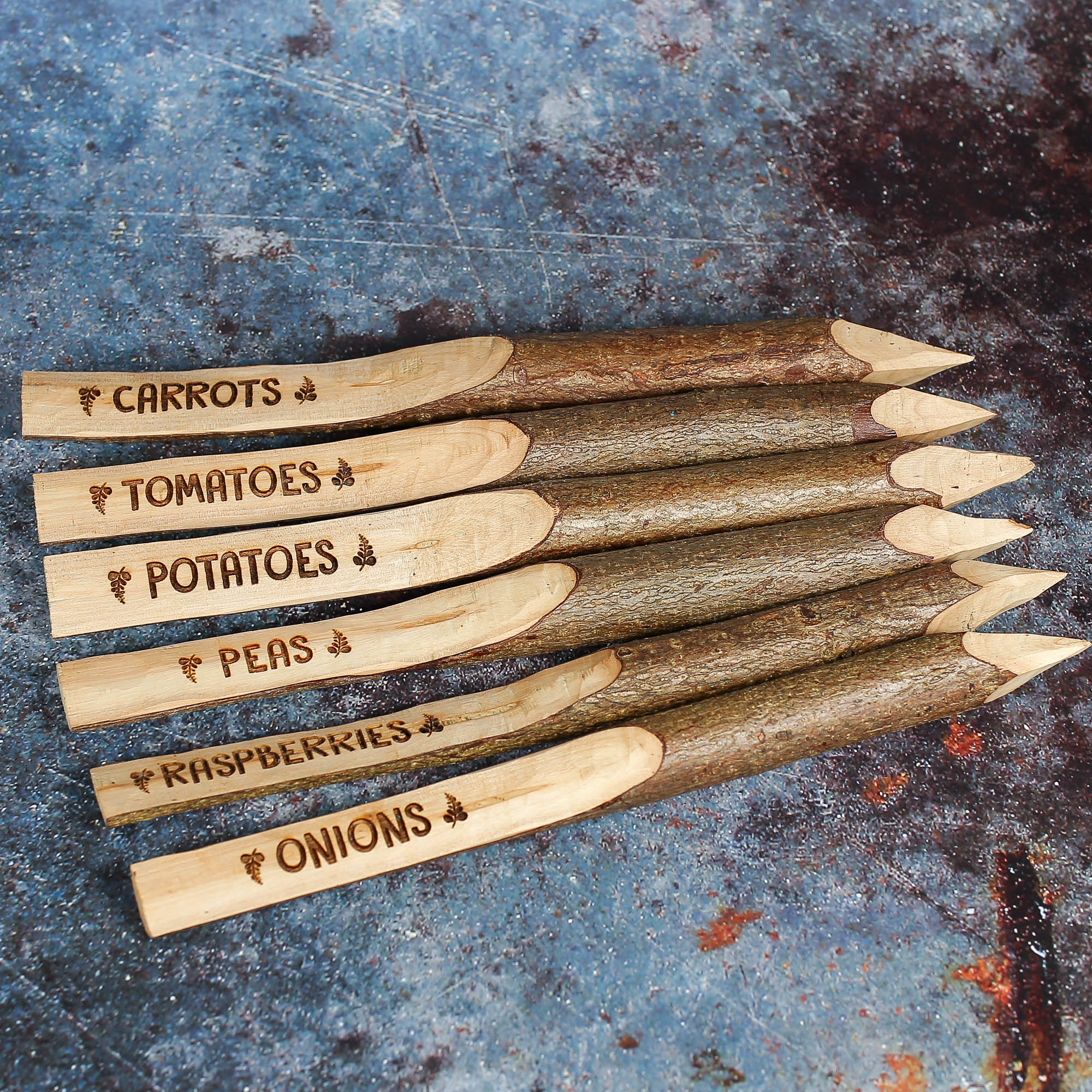 Six wooden stake like plant markers with whittled ends and engraved with vegetable names such as potatoes and carrots.
