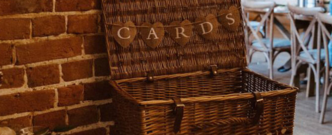 An open wicker basket on a table with a small string of bunting saying cards. In front of the basket are three more larger bunting flags with J&K engraved onto them.