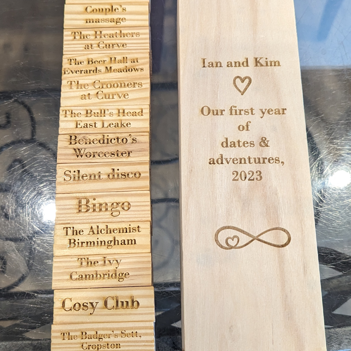 Tumble tower blocks laid out beside the holding box. The blocks are engraved with various days out whilst the box is engraved with personalisation for the couple who had enjoyed the days out. There is also heart imagery engraved onto the box.