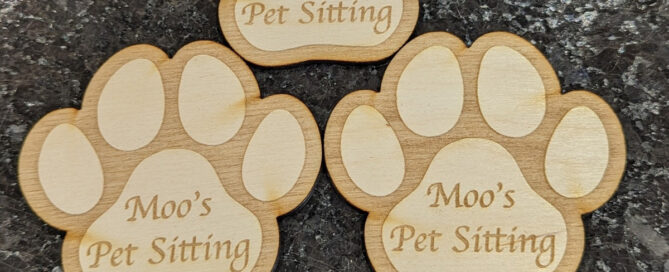Three wooden pawprints laser engraved with paw details and Moo's Pet Sitting. There is a Sharpie pen beneath to show scale - two paw prints fit across the length of the pen roughly.