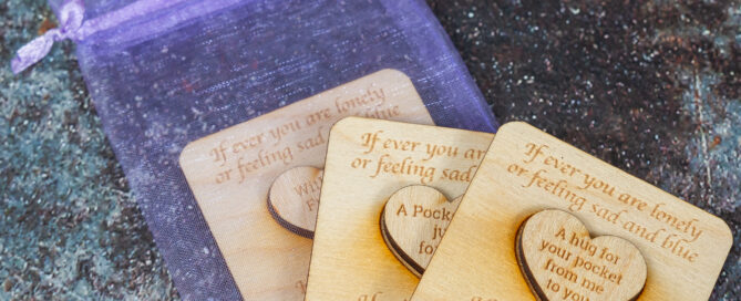 Three wooden squares on a purple organza bag. Each of the squares has a small wooden heart shape attached to them. The foremost one is laser engraved with "A hug for your pocket from me to you".